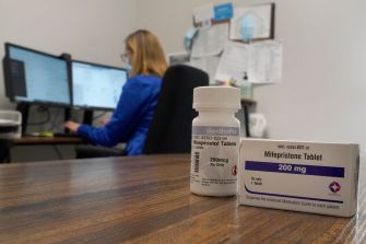A bottle of mifepristone tablets is displayed in the foreground, with a person working at a computer in the background.