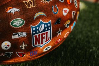 Close-up of a brown football decorated with numerous NFL team logos and stickers, resting on grass.