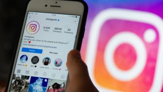 A person holds a smartphone displaying the Instagram app, with the Instagram logo blurred in the background.