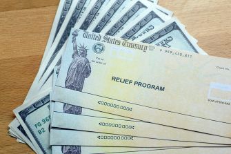 A stack of U.S. Treasury checks marked "Relief Program" alongside hundred-dollar bills on a wooden surface.
