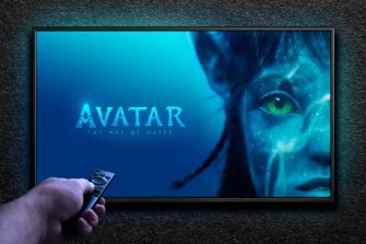 A hand holds a remote in front of a TV screen displaying Avatar: The Way of Water with a close-up blue Na’vi face.