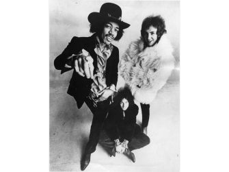 Black-and-white group portrait of a guitarist with two bandmates; one wearing a hat and pointing at the camera, the other in a fur coat.