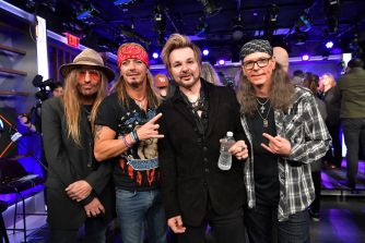 Four Mötley Crüe members pose for a backstage group photo, with cameras and purple stage lighting in the background.