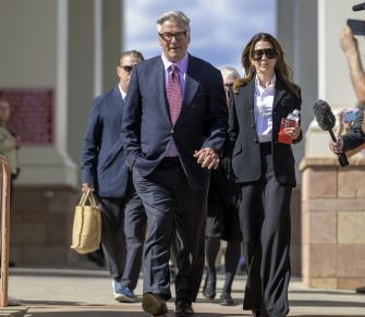 A man in a navy suit and a woman in a black pantsuit walk hand in hand down steps outside a building, surrounded by photographers and reporters.
