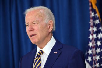 Image of a man in a suit speaking, with a blue curtain and an American flag in the background.