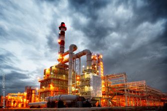 An industrial facility with pipes and smokestacks illuminated against a dramatic cloudy sky.
