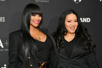 Two smiling women with long dark hair wearing black outfits pose together on a dark backdrop at a media event.