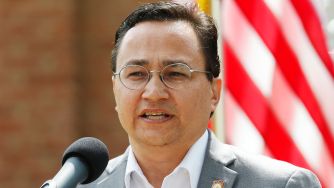 A man speaking at a microphone with an American flag in the background.