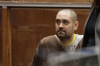 Man with a shaved head and trimmed beard sits in a courtroom behind glass during a hearing.