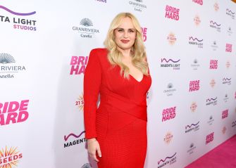 Blonde woman in a red dress posing on a pink carpet at a media event, with a backdrop covered in sponsor logos.