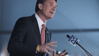 A person speaking at an event, gesturing with hands and wearing a suit and tie, with a microphone in front.