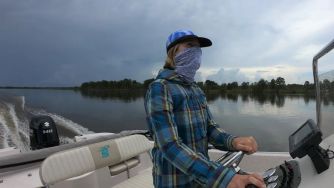 A person wearing a plaid shirt and face covering steers a small motorboat on a calm river with trees along the shoreline.