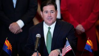 Man in a suit and green tie speaking at a podium with microphones, with American and Arizona flags in the background.