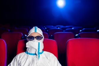 A person in a protective suit and glasses sitting alone in a movie theater with red seats and a spotlight in the background.