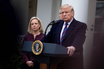 Former President Donald Trump speaking at a podium with the presidential seal, alongside a woman in a dark jacket, during a press event.