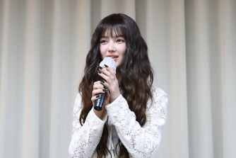 A young female singer with long wavy dark hair wearing a white lace top, holding a microphone on stage in front of curtain backdrop.