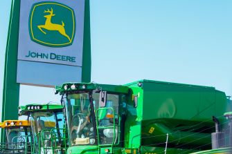 John Deere sign with yellow deer logo and green tractors parked outdoors under a blue sky