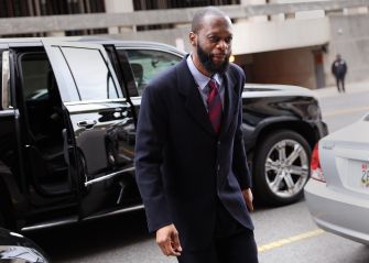 A man with a beard in a dark suit and red tie walking near parked cars outside a building.