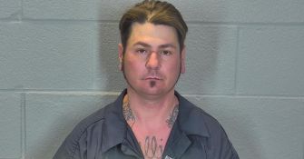 Mugshot of a tattooed man facing the camera against a gray cinder block wall.