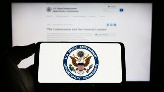 A hand holding a smartphone displaying the U.S. Equal Employment Opportunity Commission (EEOC) logo, with the agency's official website visible in the background.