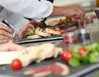 Plated foie gras being garnished in a kitchen with chefs preparing to serve.