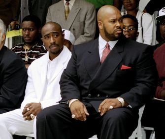 Two men in formal attire seated in the front row at a public event, one in a white suit and the other in a dark suit with a red tie, with onlookers in the background.