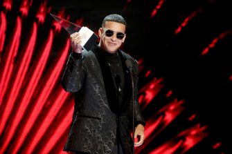 Male performer wearing sunglasses on stage, holding an award, with red stage lights in the background.