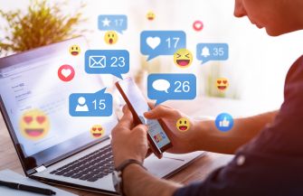 A person using a smartphone while sitting at a desk with a laptop, surrounded by social media reaction icons and notifications.