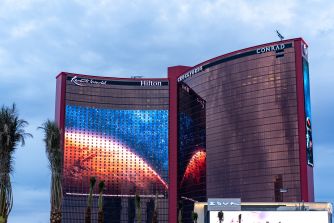 Exterior view of a Las Vegas hotel with curved glass towers, palm trees in the foreground, and signs for Conrad and Hilton on the building.