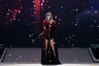 Female performer on a dark concert stage wearing a sparkling red sequin coat, singing into a microphone with a guitar at her side, with confetti in the air.