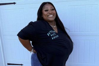 Smiling woman with long dark hair posing outdoors in front of a light blue garage door, wearing a black T-shirt and jeans.