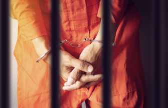A person in an orange prison outfit with handcuffs standing behind bars.