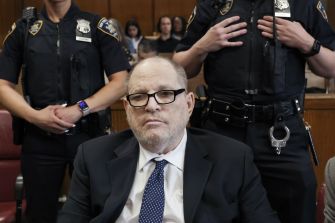 A man in a dark suit and glasses sits at a courtroom table, with uniformed officers standing behind him.