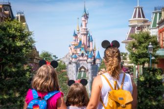 Three park visitors with backpacks walk toward Disneyland's Sleeping Beauty Castle, wearing Mickey/Minnie Mouse ears.
