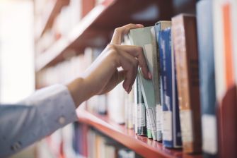 A hand reaching for a book on a library shelf filled with various books.