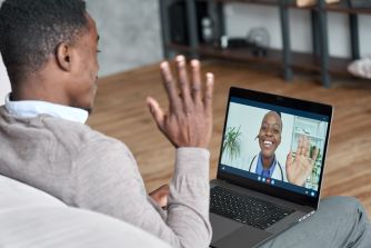 A person participating in a telehealth consultation on a laptop, smiling and waving at a healthcare provider on the screen.