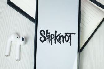 Smartphone on a desk shows the Slipknot logo on the screen, with white wireless earbuds and a notebook nearby.