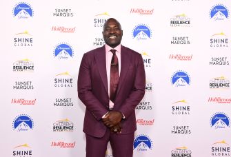 Smiling man in a burgundy suit posing on a step-and-repeat backdrop with logos for Shine Global, Paramount Pictures, Sunset Marquis, and The Hollywood Reporter.
