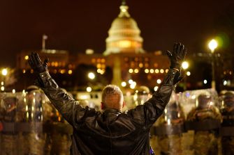 A person in a black leather jacket raises their arms in protest in front of a line of law enforcement officers, with the Capitol building illuminated in the background at night.