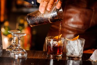 A bartender pouring a mixed drink into two glasses with garnishes at a bar.