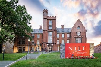 Brick NJIT campus building with a prominent red NJIT sign in the foreground, green lawn, and trees.