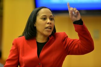 A woman in a red blazer passionately gestures while speaking, likely addressing a group or audience.