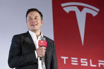 Elon Musk speaking at a Tesla event with the company logo in the background.