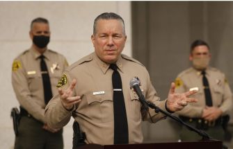 Los Angeles County Sheriff Alex Villanueva speaking at a press conference, with deputies in the background.