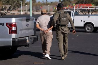 A law enforcement officer escorts an individual in handcuffs near a parked vehicle.