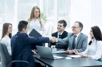 Business professionals in a boardroom; two men shake hands across a conference table as colleagues look on in a modern office setting.