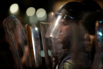 Close-up of a police officer in riot gear, wearing a helmet and clear shield, with blurred city lights in the background.