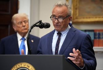 Robert F. Kennedy Jr. speaks at a podium in front of a Presidential seal, with a figure in the background.