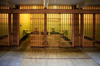 Image of an empty prison cell block with barred doors and a fluorescent light casting a yellowish glow.