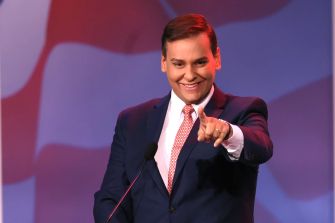 Former Rep. George Santos speaking during an event, wearing a suit and tie, with a backdrop of an American flag.